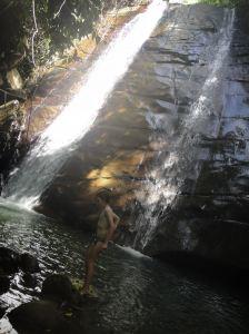 Z-in-waterfall-belaga-river-borneo-sarawak-malaysia Twin streams of water pour down a sheer brown rock face into a green pool: Z is in the foreground, preparing to dive in the water. Belaga River, Sarawak, Borneo, Malaysia.