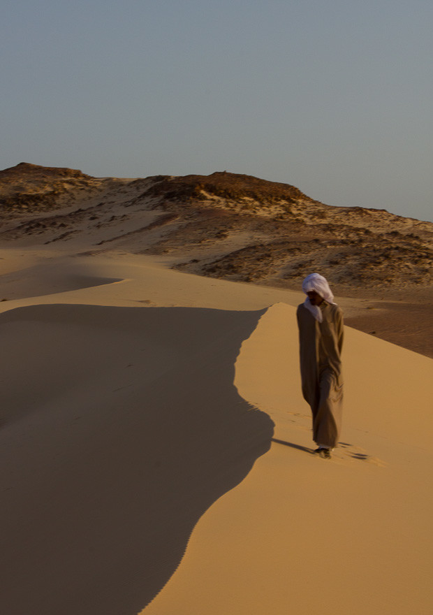 Bedouin Man Sinai Bedouin man in traditional robes and head-dress walking along a shadowed sand dune in the Sinai Desert, Egypt.