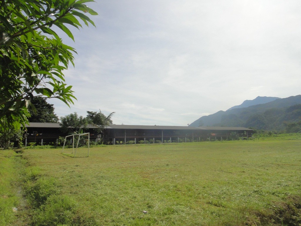 penan_longhouse_long_iman Modern wooden longhouse on grassy expanse with mountains in the background. Long Iman, Mulu, Sarawak, Borneo.