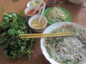 Bowl of pho with side dishes of herbs, chillis, pickled garlic, beansprouts, chilli sauce