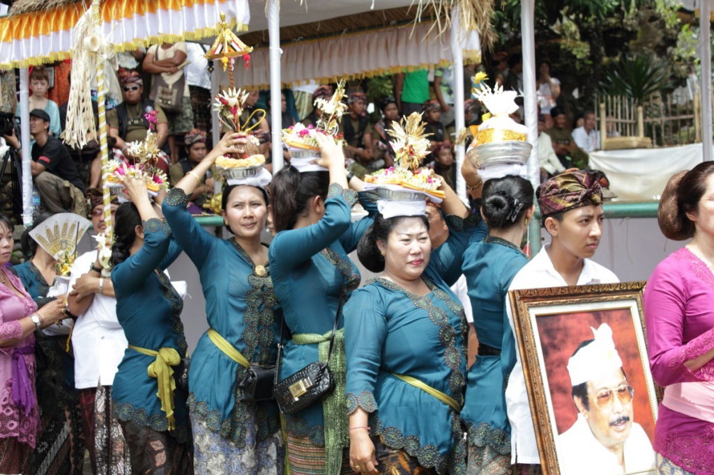 Bali cremation: women in blue blouses and gold sashes process with offerings on their heads and a portrait of the deceased.