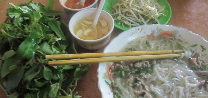 Bowl of pho with side dishes of herbs, chillis, pickled garlic, beansprouts, chilli sauce